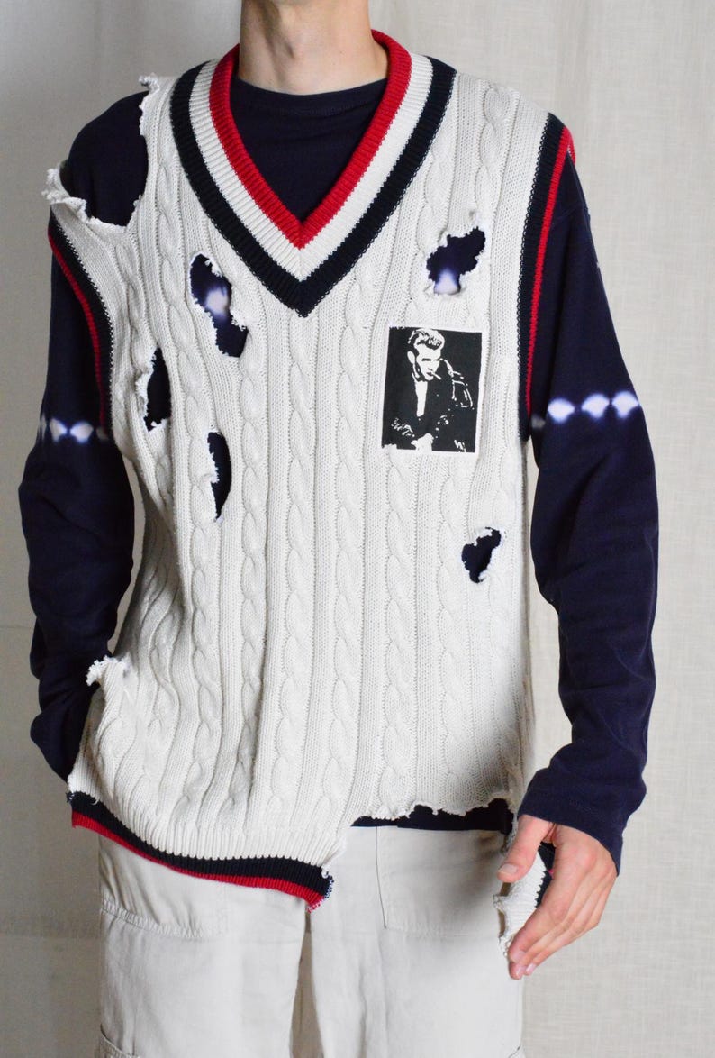 May include: Cream cable knit sweater vest with a V-neck, red and navy trim, and distressed holes. A black and white image is on the front. The model is wearing a navy blue long sleeve shirt. The vest is a unique fashion item.