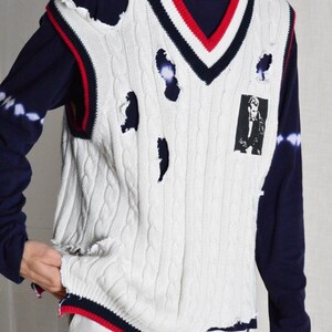 May include: Cream cable knit sweater vest with distressed detailing, red and navy trim, and a V-neck. The vest has a small black and white graphic on the front. It is worn over a navy blue long-sleeved top.