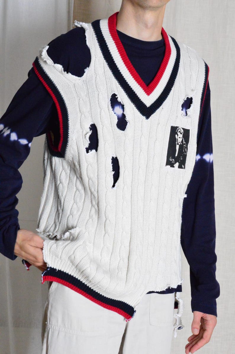 May include: Cream cable knit sweater vest with distressed detailing, red and navy trim, and a V-neck. The vest has a small black and white graphic on the front. It is worn over a navy blue long-sleeved top.