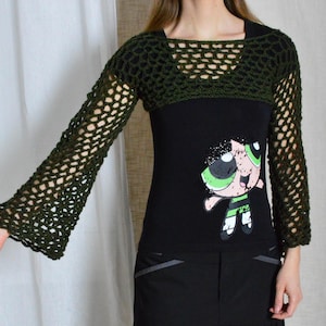 May include: A black long-sleeved shirt with a green crochet overlay. The shirt features a cartoon character with green hair and a black and white striped dress. The crochet overlay has a wide, open-weave design.