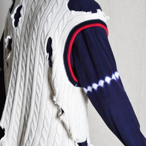 May include: Cream cable knit vest with distressed holes, navy blue and red trim. The vest is layered over a navy blue long-sleeved shirt with a white tie-dye pattern on the sleeve.