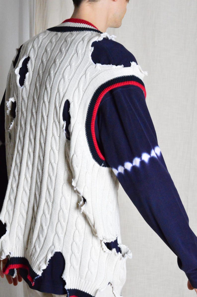 May include: Cream cable knit vest with distressed holes, navy blue and red trim. The vest is layered over a navy blue long-sleeved shirt with a white tie-dye pattern on the sleeve.