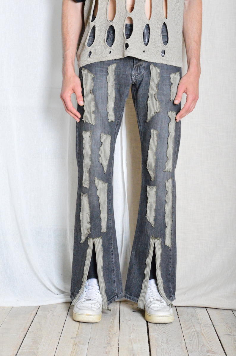 K&ouml;nnte beinhalten: Dunkelgraue Jeans mit vertikalen, strukturierten, hellgrauen Patches. Die Jeans hat ein ausgestelltes Bein-Design mit einem Schlitz am unteren Saum. Das Model tr&auml;gt wei&szlig;e Sneaker und ein hellgraues, offen gestricktes Top.