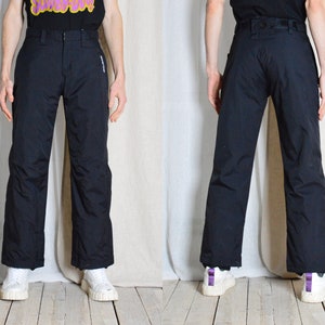 Może przedstawiać: Czarne spodnie narciarskie z kieszenią zapinaną na zamek błyskawiczny na prawej nogawce. Spodnie mają prosty krój nogawek i są wykonane z wodoodpornego materiału.