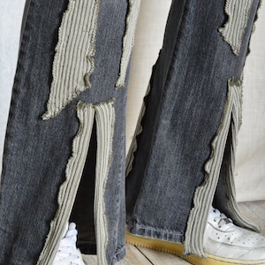 K&ouml;nnte beinhalten: Dunkelgraue Jeans mit dekorativen hellgrauen Stoffakzenten und Seitenschlitzen. Wei&szlig;e Turnschuhe sind unten zu sehen. Die Jeans haben ein einzigartiges, strukturiertes Design.