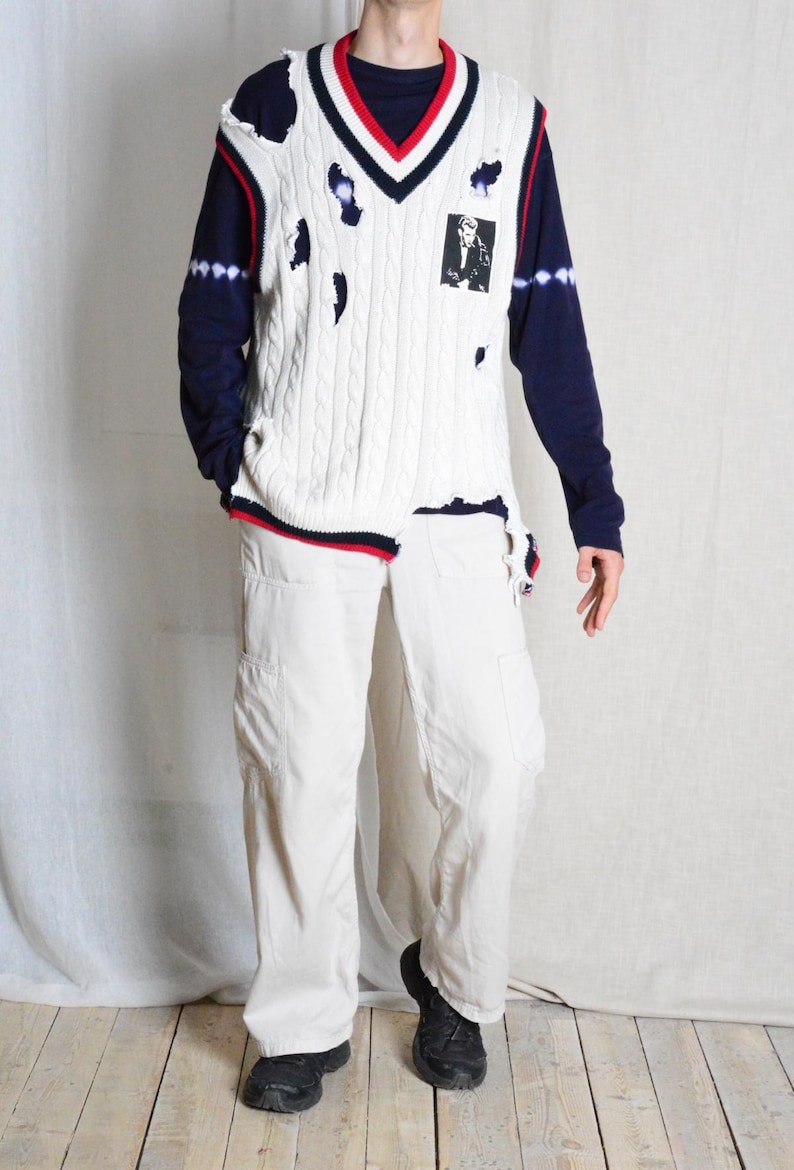 May include: A distressed white cable knit sweater vest with navy and red trim, worn over a navy long-sleeve shirt and paired with off-white cargo trousers. The vest features a black and white graphic and distressed holes.