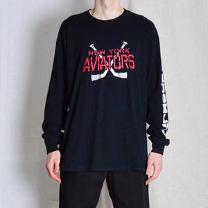 Puede incluir: Camiseta negra de manga larga con un gráfico blanco de palos de hockey cruzados y el texto "NEW YORK AVIATORS" en rojo y blanco.