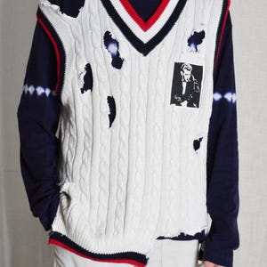 May include: Cream cable knit sweater vest with distressed holes, a V-neck, and a black and white image on the front. The vest has red and navy blue trim. The model is wearing a navy blue long sleeve shirt.