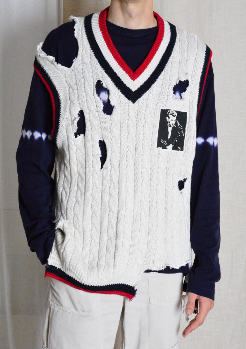 May include: Cream cable knit sweater vest with distressed holes, a V-neck, and a black and white image on the front. The vest has red and navy blue trim. The model is wearing a navy blue long sleeve shirt.