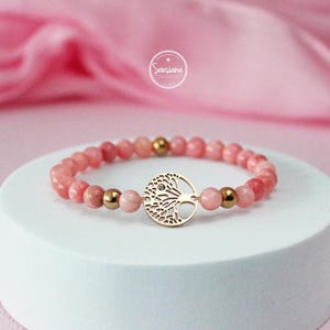 May include: A pink gemstone bracelet with a gold tree of life charm. The bracelet is made with small, round beads and has a delicate, feminine look.
