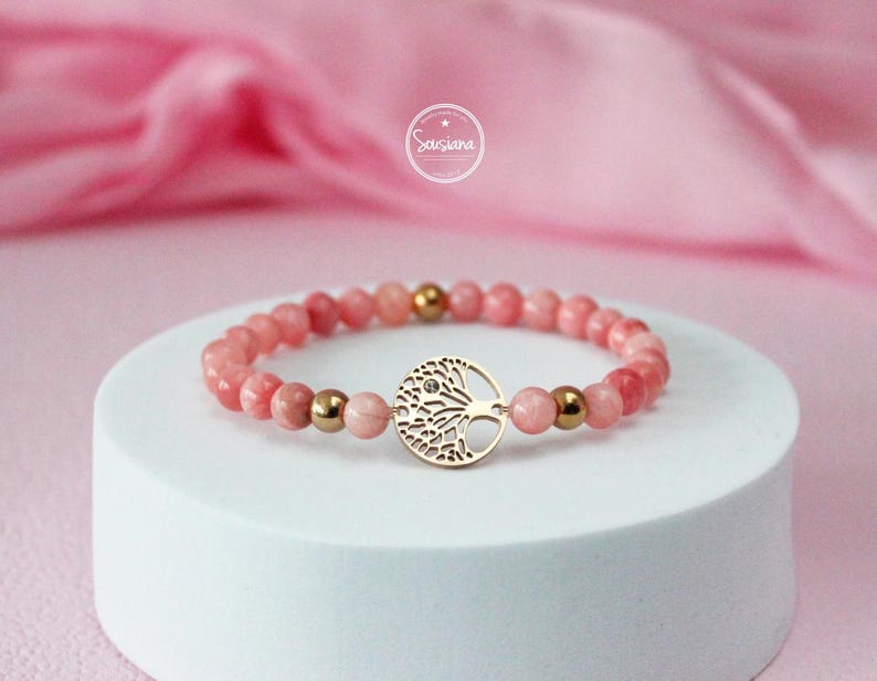 May include: A pink gemstone bracelet with a gold tree of life charm. The bracelet is made with small, round beads and has a delicate, feminine look.