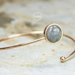 Könnte beinhalten: Ein roségoldenes Armkettchen mit einem runden, grauen Labradorit-Edelstein, der in einer goldenen Fassung sitzt. Das Armband ist offen und hat eine kleine Kugel am Ende.