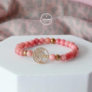 May include: A pink gemstone bracelet with a gold tree of life charm. The bracelet is made with round beads and has a gold clasp.