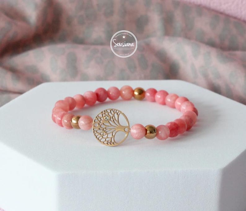May include: A pink gemstone bracelet with a gold tree of life charm. The bracelet is made with round beads and has a gold clasp.