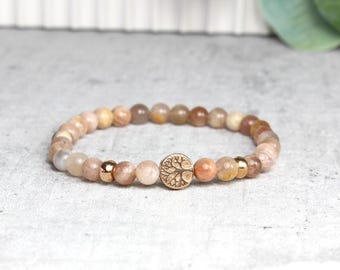 May include: A beaded bracelet with a gold tree of life charm. The beads are a mix of light brown, beige, and peach colors.