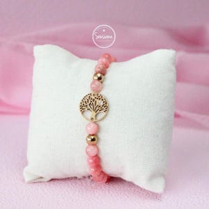 May include: A pink gemstone bracelet with a gold tree of life charm. The bracelet is on a white pillow.
