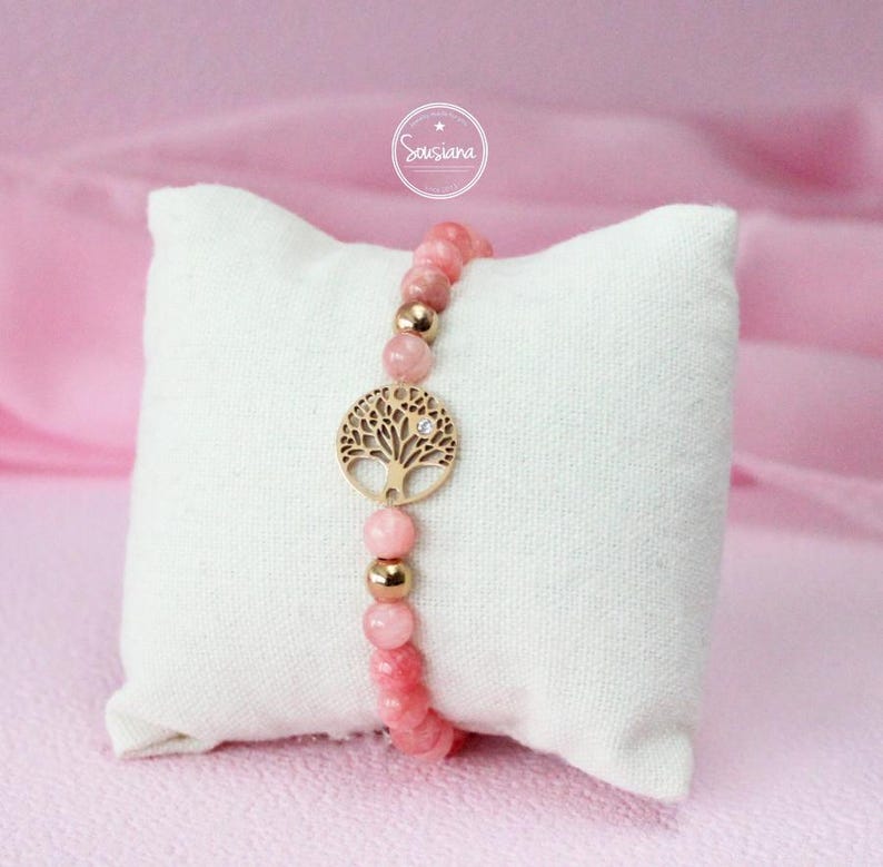 May include: A pink gemstone bracelet with a gold tree of life charm. The bracelet is on a white pillow.