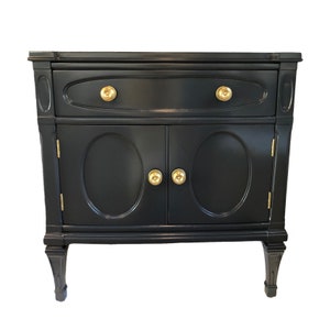 May include: A black nightstand with two doors and one drawer. The doors and drawer have oval-shaped panels and gold-colored knobs. The nightstand has four legs with a decorative design.