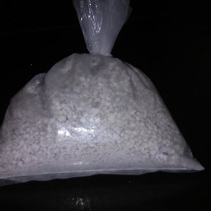 May include: A clear plastic bag filled with small, white, granular objects. The bag is tied at the top. A few of the white objects are scattered on the dark surface next to the bag.