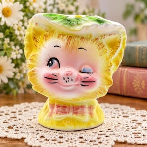 May include: A ceramic planter shaped like a winking cat head wearing a yellow bonnet with green leaf accents. The cat has yellow fur, pink cheeks, and a pink bow at the base. The facial features include black whiskers, a pink nose, and one open eye.