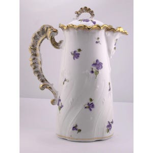 May include: White porcelain coffee pot with a lid, adorned with purple floral designs and gold trim. The handle and lid detail feature ornate gold accents. A decorative piece for serving coffee or tea.