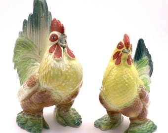 May include: Two ceramic chicken figurines featuring yellow chests, brown wings, and green tail feathers. Each bird has a red comb and wattle, with detailed textured plumage and green feet, standing upright in a pair on a plain white background.