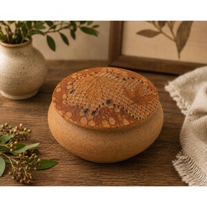 May include: A round, tan ceramic lidded jar with a textured, matte finish. The lid features a detailed, dark brown embossed pattern of maple leaves and a delicate, net-like lace design etched into the surface of the clay material.