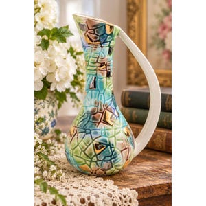 May include: A ceramic pitcher with a tall, slender neck and a wide, rounded base. The surface features a mosaic-like pattern of turquoise, blue, and brown segments with gold-toned accents. It has a single, smooth, white curved handle.