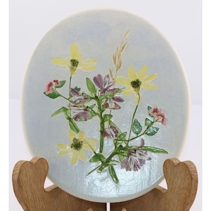May include: A round, light blue ceramic plate featuring a floral design. The flowers are yellow, purple, and pink, with green leaves and a tan stalk. The plate is displayed on a wooden stand.