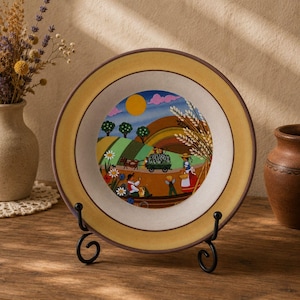 May include: A decorative ceramic plate with a yellow rim and a painted pastoral scene. The scene depicts a sunny day with rolling hills, trees, and figures harvesting crops. The plate is displayed on a black metal stand.