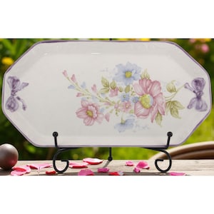 May include: White rectangular serving platter with a floral design in pink, blue, and green. The platter has a purple border and bow accents. It is displayed on a black metal stand, with rose petals scattered around.