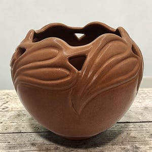 May include: A round, terracotta-colored ceramic bowl featuring a smooth, matte finish. The upper rim is scalloped with cut-out triangular openings between raised, leaf-like relief patterns that wrap around the exterior of the vessel.