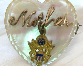 May include: A white heart-shaped pin with a gold inscription that reads "Mother". A gold charm with an American eagle and shield hangs from the inscription.