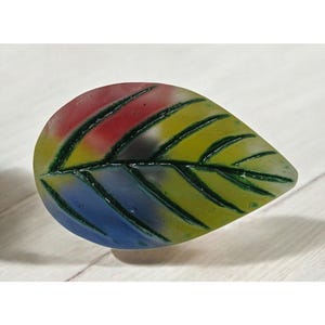 May include: A colorful glass leaf with green veins and a translucent design. The leaf is a vibrant mix of red, yellow, and blue.
