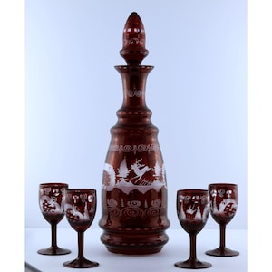 May include: A deep red glass decanter with a matching stopper and four small stemmed glasses. The set features white etched designs of deer, pine trees, and swirling patterns on the surface of the glass, creating a contrast with the red color.