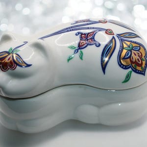 May include: A white porcelain trinket box shaped like a sleeping cat. The box is decorated with a colorful floral design in shades of blue, red, gold, and green.