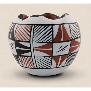 May include: A round, white ceramic bowl with a scalloped rim. The bowl is decorated with a geometric pattern of black, red, and white shapes. The design includes triangles, stripes, and squares, creating a visually striking aesthetic.