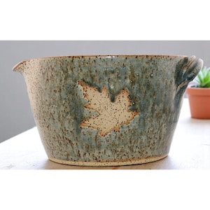 May include: A ceramic bowl with a blue-green glaze and a beige leaf design. The bowl has a spout and a handle. The speckled glaze gives the bowl a rustic appearance. The bowl is suitable for serving or as a decorative piece.