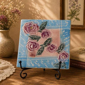 May include: Square ceramic tile with a hand-painted floral design. The tile features pink roses with green leaves against a blue and pink background. It is displayed on a black metal stand, showcasing a decorative art piece.