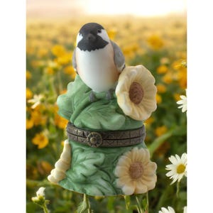 May include: A porcelain trinket box with a detailed design of a black-capped chickadee perched on a green leaf with yellow flowers. The box has a hinged lid and a brass clasp.