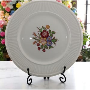 May include: White ceramic plate with a floral bouquet design in the center. The bouquet features red, yellow, blue, and green flowers. The plate has a ribbed border and is displayed on a black metal stand.