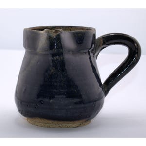 May include: A handmade, dark blue ceramic pitcher with a rounded body and a curved handle. The pitcher has a spout for pouring and a textured, unglazed base. The glaze has a glossy finish.