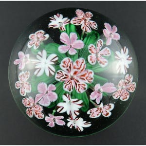 May include: A spherical glass paperweight featuring a clear dome encasing a floral arrangement. The design includes multiple pink and white flowers with dark red veining, surrounded by green glass leaves, all set against a dark background.