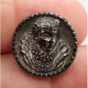 May include: A detailed, antique silver button with a raised relief of a regal figure. The button features a detailed face, ornate collar, and decorative border. The button is round and has a slightly tarnished appearance, suggesting age.