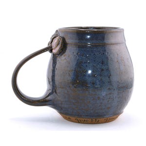 May include: A handmade ceramic mug with a dark blue glaze and a brown rim. The mug has a large handle and a decorative element where the handle attaches. The base of the mug has handwritten text. A unique piece for enjoying beverages.