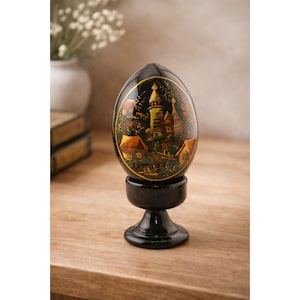 May include: A decorative black egg with a painted landscape scene, set on a matching black stand. The egg features a detailed depiction of buildings with gold accents. The stand has a pedestal base. The egg is a unique decorative item.