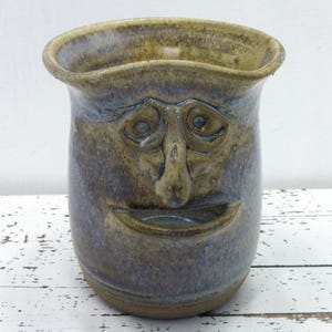 May include: A ceramic mug featuring a sculpted human face on the front. The vessel has a mottled glaze in shades of blue, brown, and tan. It has a wide, flared rim and a deep, carved mouth opening. The surface texture is smooth and glossy.