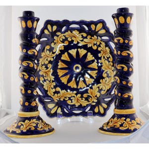 May include: Two tall, spiral-shaped ceramic candlesticks in deep blue with yellow floral accents, flanking a matching square ceramic plate with a central sunburst pattern and an intricate, open-work scalloped border in blue and yellow.