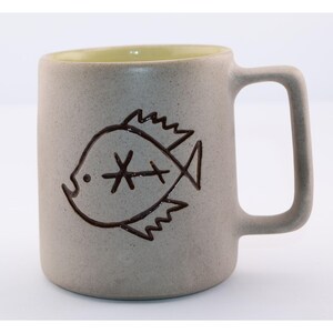 May include: A ceramic mug with a light gray exterior and a yellow interior. A dark brown, stylized fish design is etched on the side. The mug has a large, rectangular handle.