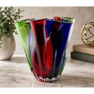 May include: A colorful glass vase with a unique, wavy design. The vase features vibrant sections of green, red, and blue glass. The vase is a decorative piece for home decor.
